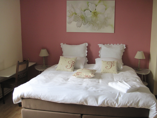 Tweepersoonsbed met witte lakens en kussens, roze muur, nachtkastjes en lampen, bloemenfoto boven het bed.
